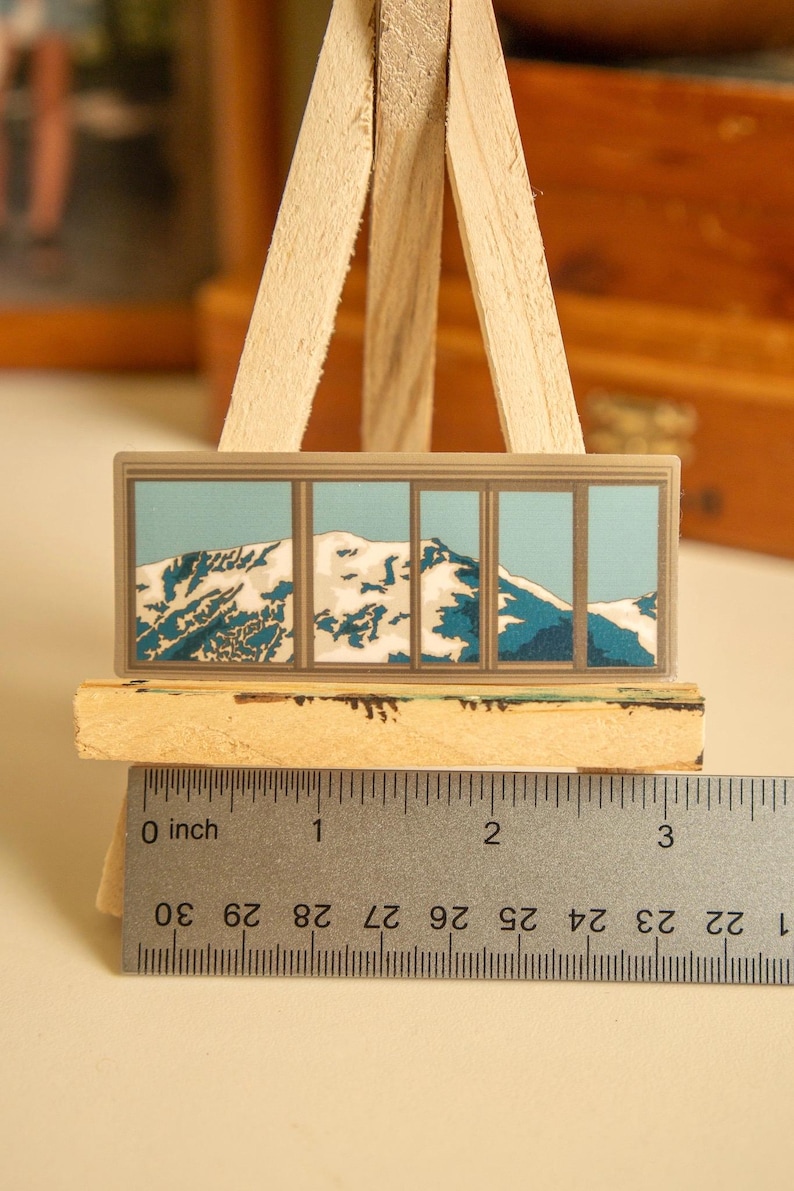 May include: A sticker of a mountain range with snow-capped peaks, viewed through a window with multiple panes. The sticker is approximately 2 inches wide and is displayed on a wooden easel.