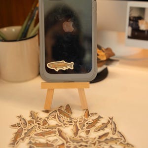 May include: A gray phone case with a fish sticker on a small wooden easel, surrounded by fish stickers. The stickers feature a detailed illustration of a fish with brown, orange, and gray tones. The phone case has a clear back and a gray border.