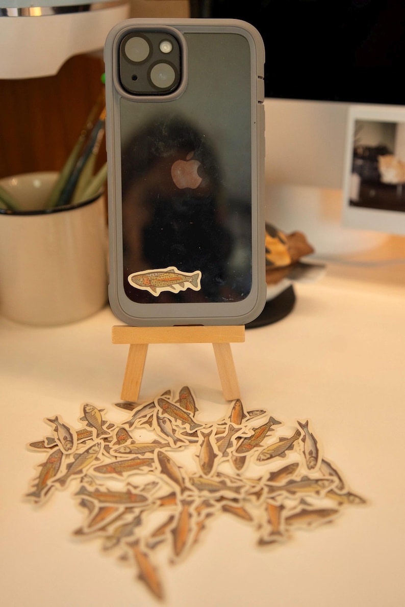 May include: A gray phone case with a fish sticker on a small wooden easel, surrounded by fish stickers. The stickers feature a detailed illustration of a fish with brown, orange, and gray tones. The phone case has a clear back and a gray border.