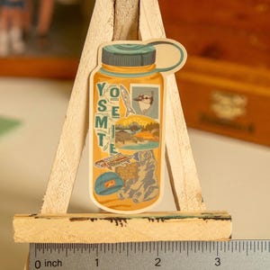 May include: A yellow and blue sticker of a water bottle with the word "Yosemite" printed vertically on the side. The sticker has a collage of images including a bird, fish, and a bear.