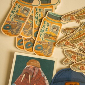 May include: A collection of stickers featuring a walrus wearing a hat, a blue beanie with a mountain graphic, and several fish and water bottle stickers with the text "Yosemite" on them.
