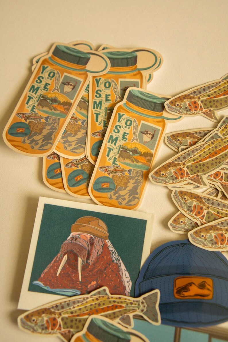 May include: A collection of stickers featuring a walrus wearing a hat, a blue beanie with a mountain graphic, and several fish and water bottle stickers with the text "Yosemite" on them.