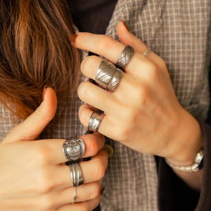 May include: A hand wearing several silver rings with different designs. The rings are worn on the index, middle, ring, and pinky fingers.