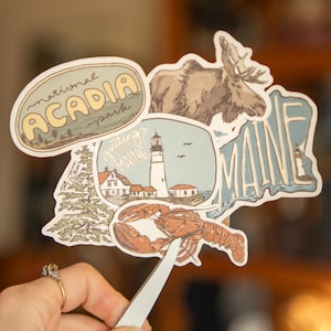 May include: A selection of stickers with a Maine theme, including images of a moose, a lighthouse, a lobster, and the words "Acadia National Park" and "Maine". The stickers have a vintage, hand-drawn aesthetic with a beige, blue, and brown colour scheme.