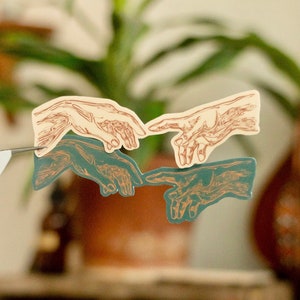 May include: Two sets of hands reaching towards each other, one set in cream with brown lines and the other in teal with brown lines. The hands are in a stylized design reminiscent of the famous Renaissance painting.