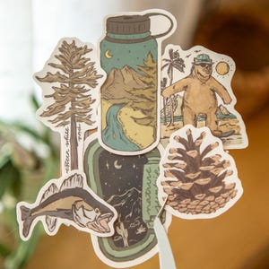 May include: A collection of nature-themed stickers featuring illustrations of a pine tree, a fish, a bear at the beach, a pine cone, and a water bottle with a landscape design. The stickers have a vintage, hand-drawn aesthetic.