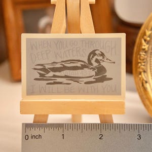 Op de afbeelding: Een kleine houten ezel draagt een rechthoekig bord met een eendenillustratie en de tekst "WHEN YOU GO THROUGH DEEP WATERS I WILL BE WITH YOU." Het bord is ongeveer 5 cm breed. Een liniaal staat onderaan.