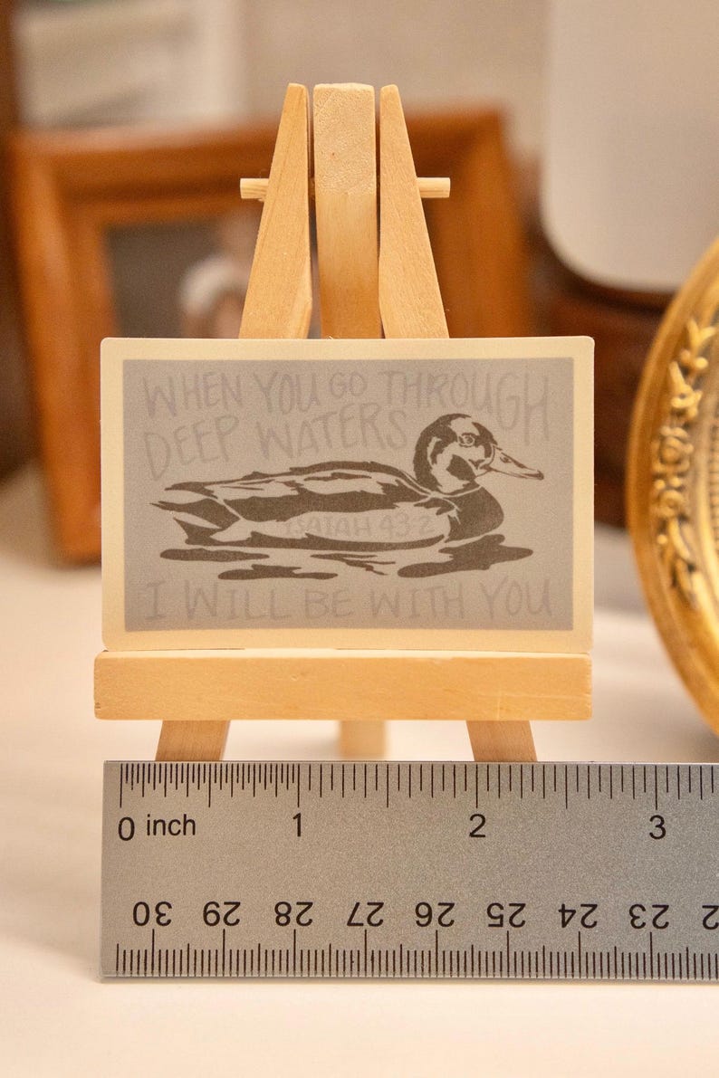 Op de afbeelding: Een kleine houten ezel draagt een rechthoekig bord met een eendenillustratie en de tekst "WHEN YOU GO THROUGH DEEP WATERS I WILL BE WITH YOU." Het bord is ongeveer 5 cm breed. Een liniaal staat onderaan.