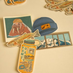 May include: A collection of stickers featuring a variety of designs, including a walrus wearing a hat, a fish, a mountain range, and a water bottle with the word "Yosemite" on it. The stickers are on a white background.
