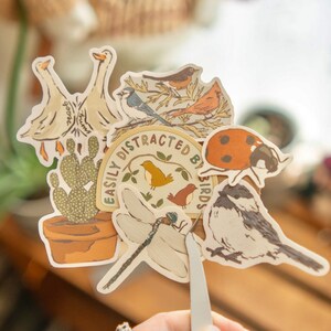 Peut inclure: Un assortiment d'autocollants avec des illustrations d'oiseaux, une coccinelle, un cactus et une libellule. Un autocollant porte l'inscription "Easily Distracted By Birds". Les autocollants sont tenus par une pince.