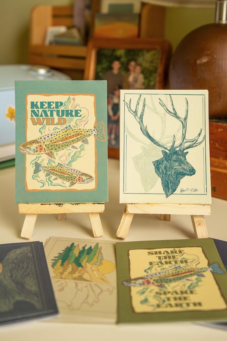 May include: Four illustrated cards with nature-themed designs. The cards are on small wooden easels. The cards feature a trout, an elk, a mountain scene, and a trout with the text "Share the Earth, Spare the Earth".