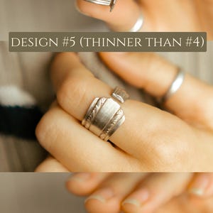 May include: A silver ring made from a repurposed spoon. The ring is open and has a decorative design on the band. The ring is shown on a person's finger.