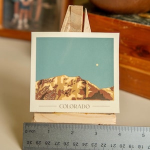 May include: A vintage-style postcard featuring a mountain range with a light blue sky and a small white moon. The postcard has the word "COLORADO" printed at the bottom.