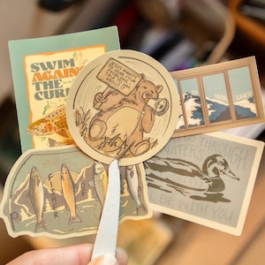 Op de afbeelding: Een verzameling stickers met diverse ontwerpen. E&eacute;n sticker toont een beer met een bord met de tekst "JESUS WANTS TO HEAR YOUR PROBLEMS - TALK TO HIM ABOUT IT". Andere stickers tonen vissen, een eend en een berglandschap.