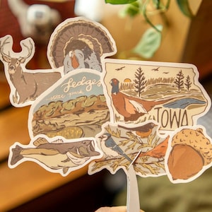 Puede incluir: Una colección de pegatinas con temática de Iowa que presentan ilustraciones de un ciervo, un pavo, un faisán, un pez, pájaros y una bellota. Las pegatinas tienen un estilo vintage, dibujado a mano, con colores tierra. Una pegatina dice "Ledges State Park" y otra "Iowa".