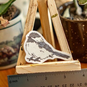 Peut inclure: Un autocollant d'un oiseau m&eacute;sange est pr&eacute;sent&eacute; sur un petit chevalet en bois. L'oiseau est blanc avec des marques grises et noires. Une r&egrave;gle m&eacute;tallique avec des mesures en pouces est plac&eacute;e devant le chevalet.