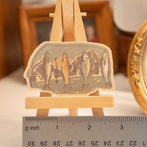 Op de afbeelding: Een sticker met vijf vissen in verschillende kleuren tegen een berglandschap. De sticker wordt weergegeven op een kleine houten ezel met een liniaal in inches. Het woord "FISH" is gedeeltelijk zichtbaar.