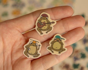 May include: Three frog stickers with different expressions. One frog is playing a guitar, another is wearing a hat and holding a cup, and the third is wearing a hat and has a surprised expression.