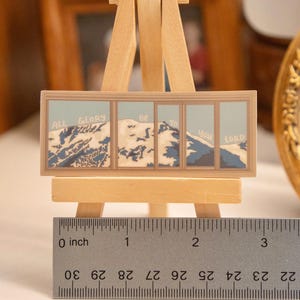 Op de afbeelding: Een miniatuurschilderij op een kleine houten ezel. Het kunstwerk toont een berglandschap met de woorden "ALL GLORY BE TO YOU LORD" in een blauw-wit kleurenschema. Het schilderij is ongeveer 5 cm breed.