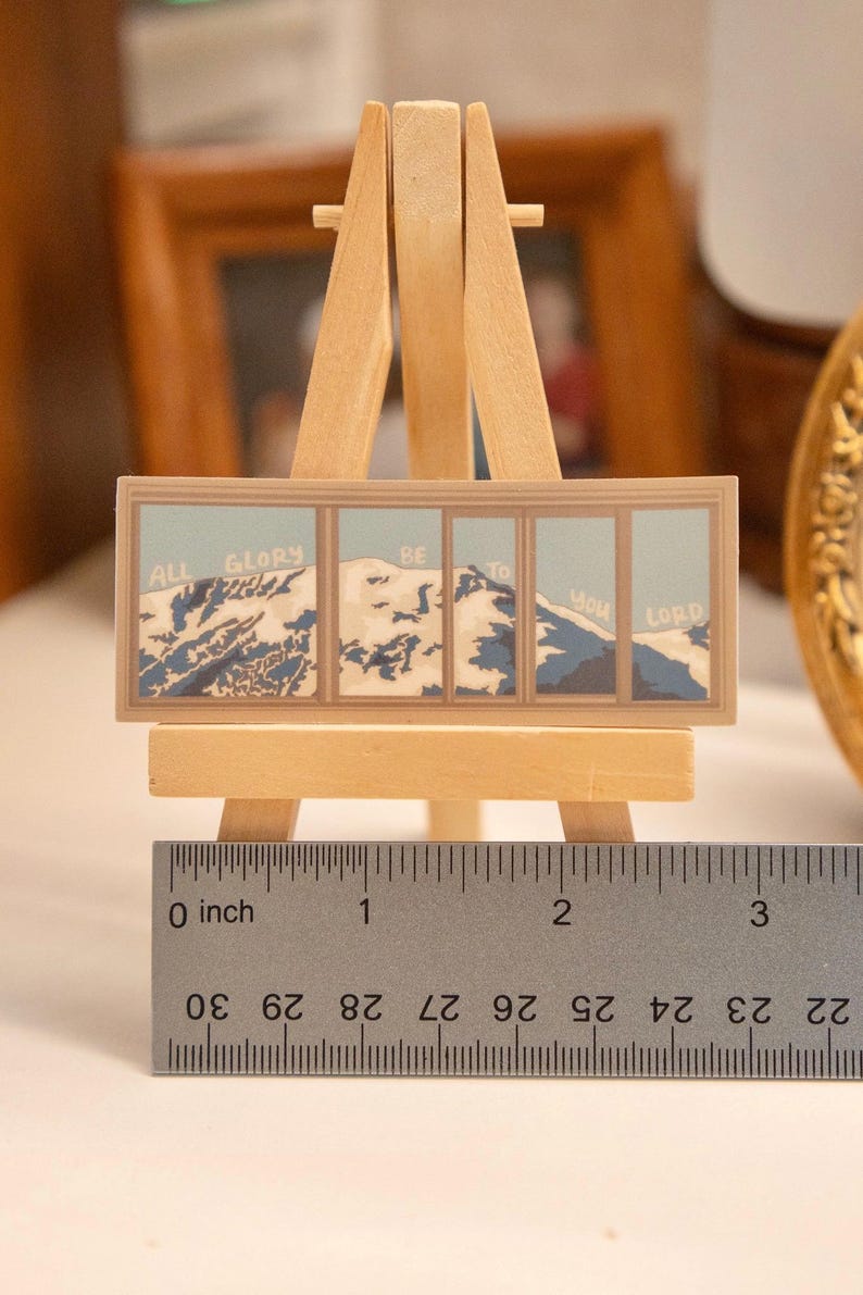 Op de afbeelding: Een miniatuurschilderij op een kleine houten ezel. Het kunstwerk toont een berglandschap met de woorden "ALL GLORY BE TO YOU LORD" in een blauw-wit kleurenschema. Het schilderij is ongeveer 5 cm breed.