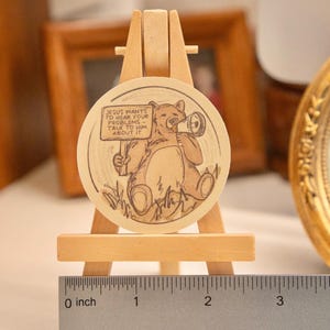 Op de afbeelding: Een rond houten kunstwerk met een beer die een megafoon vasthoudt en een bord met de tekst "JESUS WANTS TO HEAR YOUR PROBLEMS - TALK TO HIM ABOUT IT." Het wordt tentoongesteld op een kleine houten ezel. De diameter is ongeveer 5 cm.