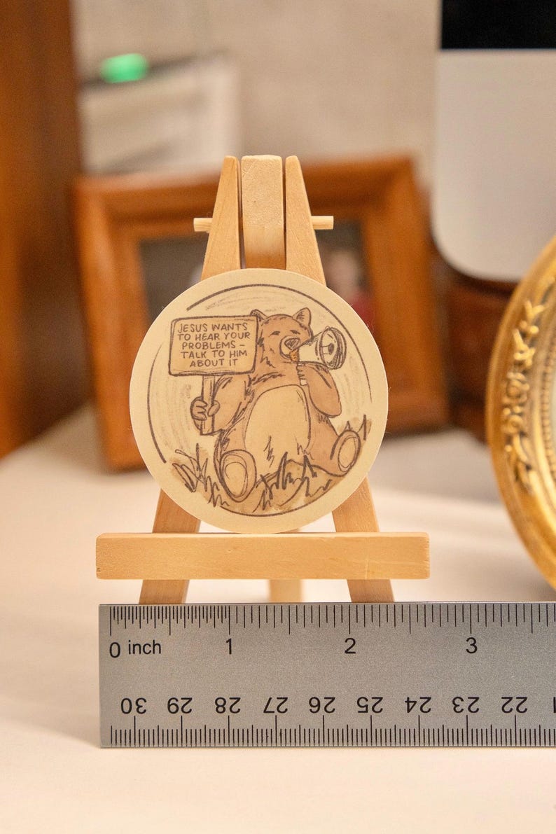 Op de afbeelding: Een rond houten kunstwerk met een beer die een megafoon vasthoudt en een bord met de tekst "JESUS WANTS TO HEAR YOUR PROBLEMS - TALK TO HIM ABOUT IT." Het wordt tentoongesteld op een kleine houten ezel. De diameter is ongeveer 5 cm.