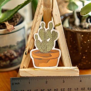 Peut inclure: Un autocollant repr&eacute;sentant un cactus dans un pot en terre cuite, pr&eacute;sent&eacute; sur un petit chevalet en bois. Le cactus est vert avec des points blancs. Une r&egrave;gle indique que l'autocollant mesure environ 5 cm de large.