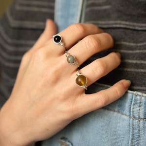 May include: Three silver wire wrapped rings with different colored stones. The ring on the index finger has a black stone, the middle finger has a gray stone, and the ring finger has a yellow stone.
