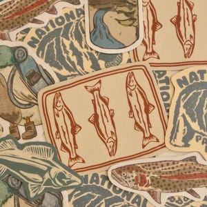 Peut inclure: Une collection d'autocollants assortis sur le thème de la nature, incluant des illustrations de truites tachetées, de saumons dans un cadre rectangulaire, de paysages montagneux et d'ours, dans une palette de couleurs bleu, rouge et beige.