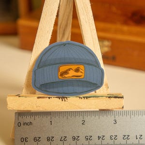 May include: A blue beanie with a small orange mountain graphic patch. The beanie is about 2 inches in diameter.