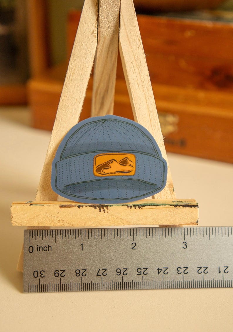 May include: A blue beanie with a small orange mountain graphic patch. The beanie is about 2 inches in diameter.