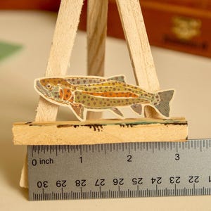 May include: A colorful sticker of a trout fish on a wooden easel with a ruler measuring 3 inches. The fish is brown, orange, and green with black spots.