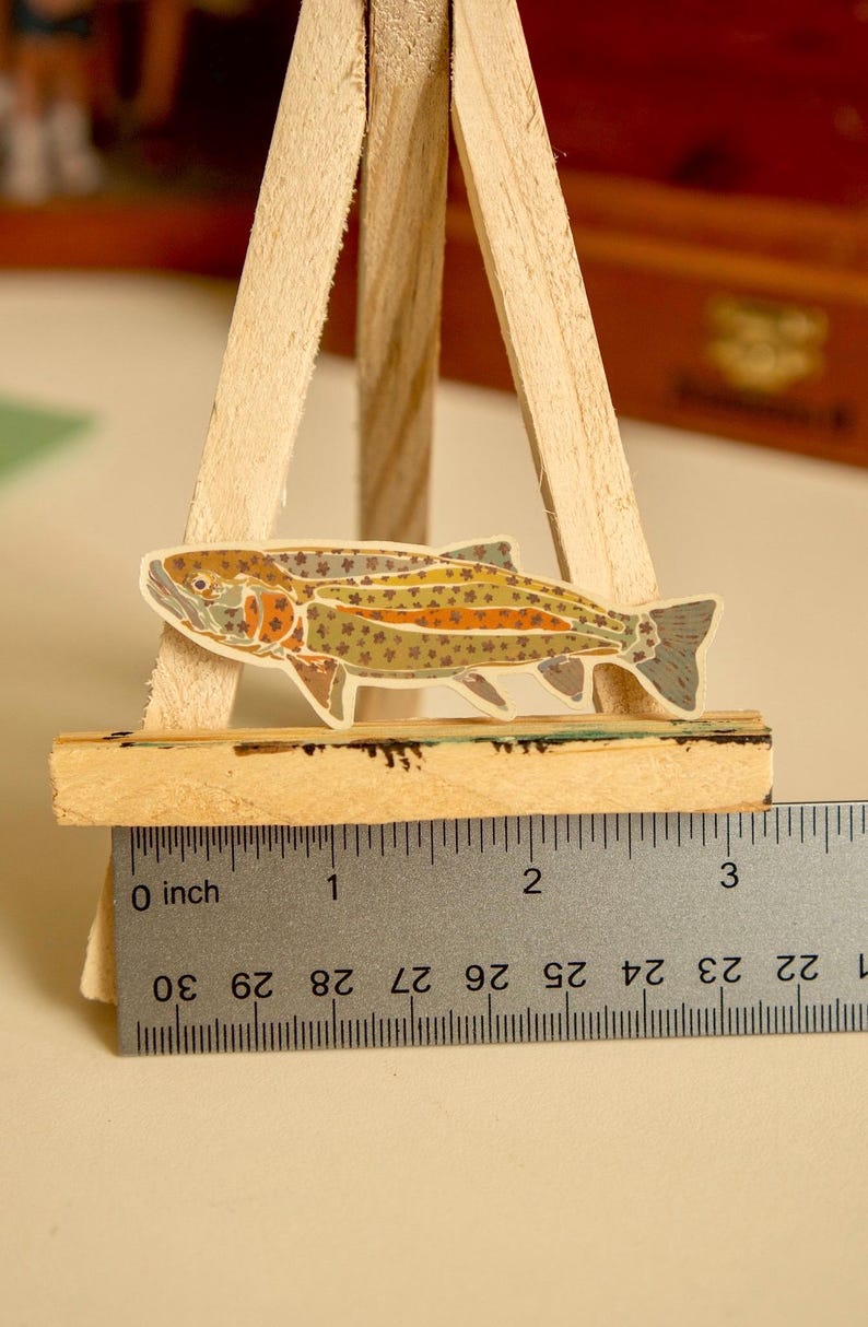 May include: A colorful sticker of a trout fish on a wooden easel with a ruler measuring 3 inches. The fish is brown, orange, and green with black spots.