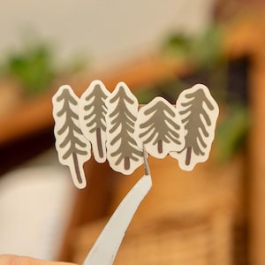 May include: A cluster of four tree-shaped stickers with a white border and a gray-green fill. The stickers are being held by a pair of white tweezers. The background is blurred, with hints of green and brown.