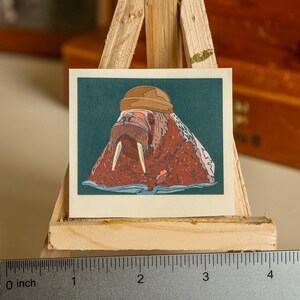 May include: A sticker featuring a cartoon walrus wearing a brown hat. The walrus is brown and pink with white tusks. The background is teal.