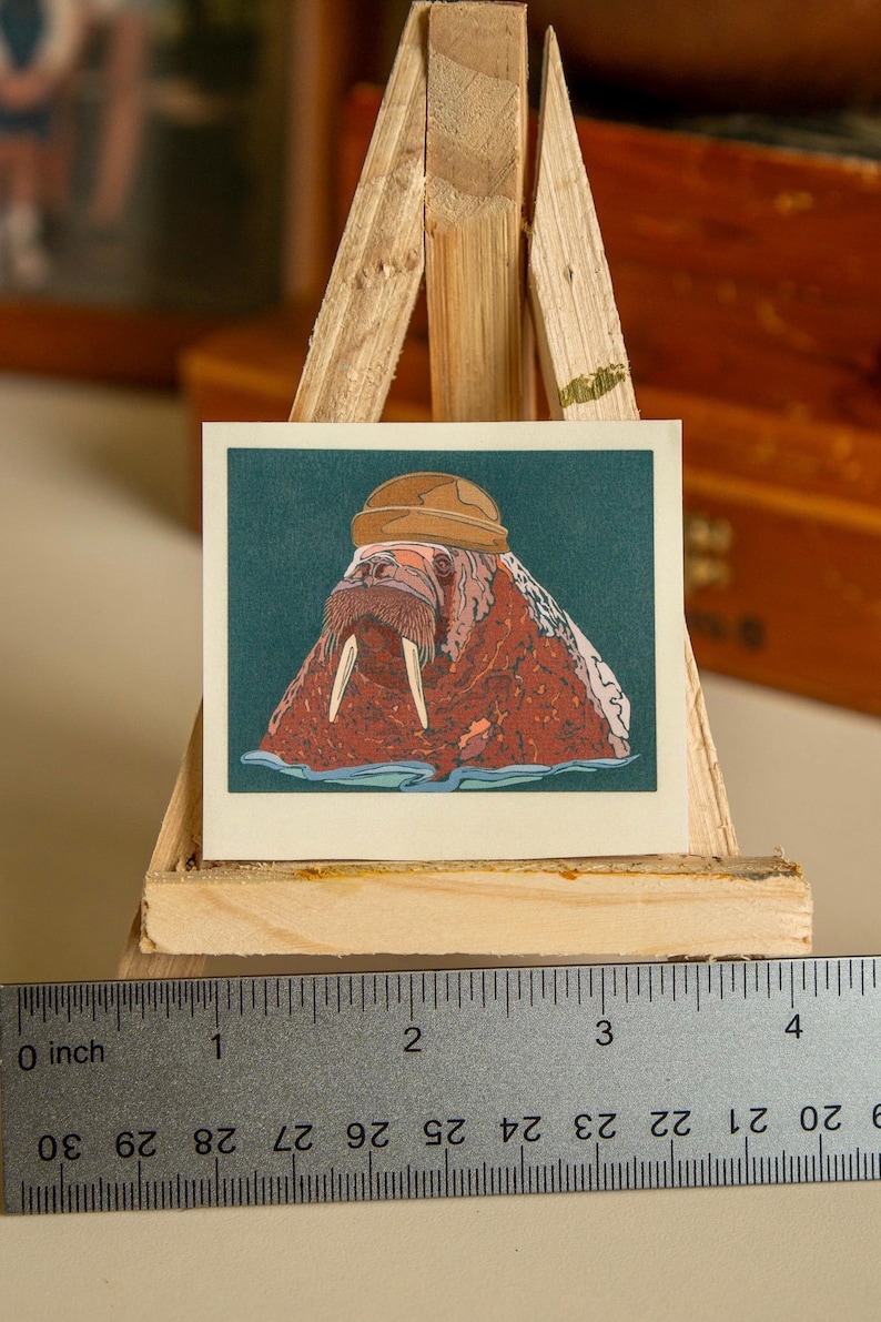 May include: A sticker featuring a cartoon walrus wearing a brown hat. The walrus is brown and pink with white tusks. The background is teal.