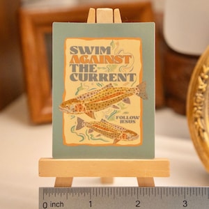 Op de afbeelding: Een kleine, rechthoekige kunstdruk op een miniatuur houten ezel. De print toont twee visillustraties en de tekst "SWIM AGAINST THE CURRENT" en "FOLLOW JESUS" in een vintage stijl. De print is ongeveer 5 cm breed.