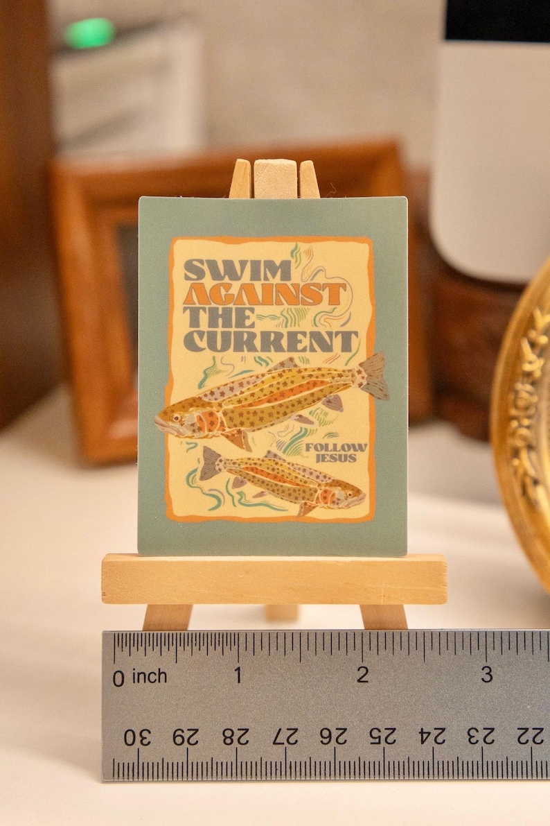 Op de afbeelding: Een kleine, rechthoekige kunstdruk op een miniatuur houten ezel. De print toont twee visillustraties en de tekst "SWIM AGAINST THE CURRENT" en "FOLLOW JESUS" in een vintage stijl. De print is ongeveer 5 cm breed.