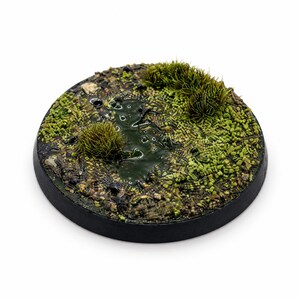 May include: A circular miniature display base featuring a textured terrain surface with dark brown soil, small pebbles, patches of bright green moss, and two tufts of tall grass. A glossy, dark green puddle effect occupies the centre area.