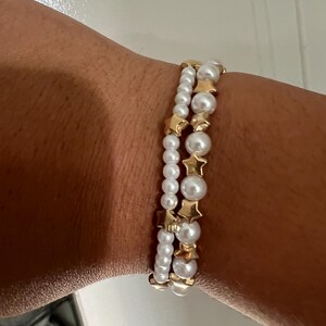 Puede incluir: Tres pulseras apiladas en una muñeca. Las pulseras presentan perlas blancas y cuentas doradas en forma de estrella. Las perlas varían en tamaño y las pulseras están unidas con elástico. Un accesorio de moda.