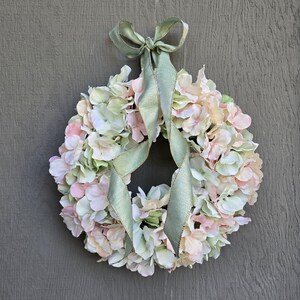 May include: A wreath made of pink and white hydrangeas with a sage green ribbon.