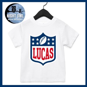 May include: White t-shirt with a blue and red football logo. The logo has stars and the name "LUCAS" in red.