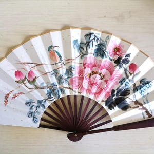 Könnte beinhalten: Handbemalter japanischer Fächer mit einem Vogel, rosa Blüten und grünen Blättern. Der Fächer hat einen dunkelbraunen Holzgriff und einen cremefarbenen Hintergrund mit vertikalen Streifen. Das Design ist japanisch inspiriert.