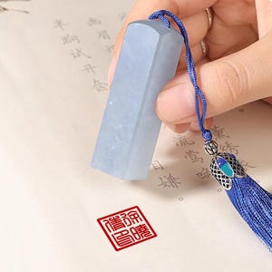 Könnte beinhalten: Ein hellblaues, rechteckiges Jade-Siegel mit einer blauen Kordel und Quaste. Das Siegel wird in einer Hand gehalten, und ein roter quadratischer Stempel ist auf dem Papier sichtbar. Die Quaste hat ein dekoratives Metallstück.