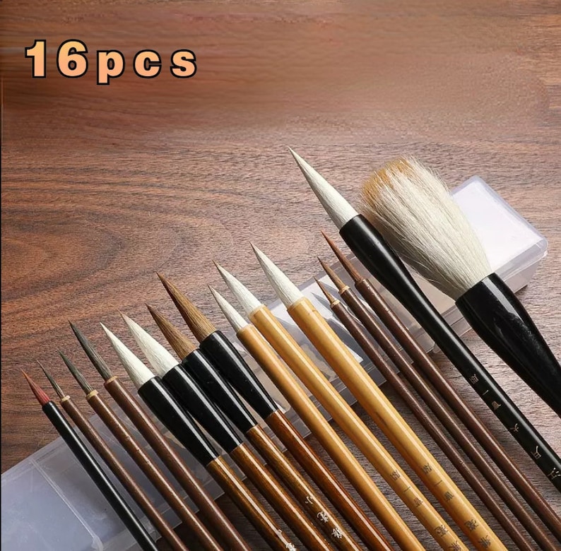 K&ouml;nnte beinhalten: Ein Set mit 16 Kalligraphie-Pinseln in einem transparenten Kunststoff-Etui. Die Pinsel haben schwarze und braune Griffe und wei&szlig;e Borsten. Die Pinsel sind f&auml;cherf&ouml;rmig angeordnet, wobei der gr&ouml;&szlig;te Pinsel oben und der kleinste unten ist.