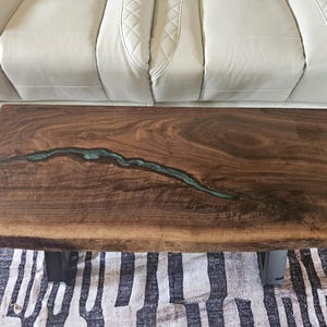 May include: A rectangular coffee table featuring a dark brown natural wood slab with a live edge. A long, narrow, irregular fissure running through the center of the wood is filled with a translucent, shimmering teal-colored epoxy resin.