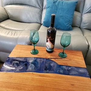 May include: A wooden coffee table with a blue and white epoxy river design. Two wine glasses and a bottle of wine are on the table.