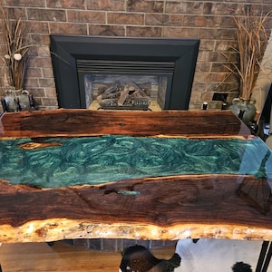 May include: A rectangular live edge wood table featuring a central river design made of translucent teal and green epoxy resin with swirling patterns. The table has a polished, high-gloss finish and is supported by four black metal hairpin legs.