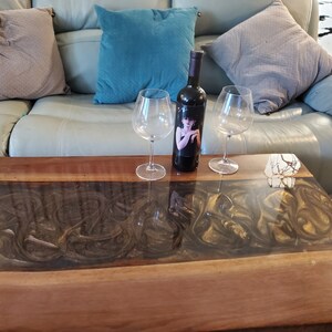 May include: A rectangular coffee table featuring a natural wood edge and a central resin inlay with swirling dark brown and gold patterns. A dark wine bottle with a label depicting a person in a black dress stands between two clear glass wine glasses.