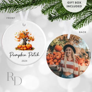 May include: A white ceramic ornament with a watercolor illustration of a fall tree and pumpkins. The text "Pumpkin Patch 2024" is written below the illustration. The back of the ornament is a photo of a woman in a striped shirt standing in front of a pile of pumpkins.